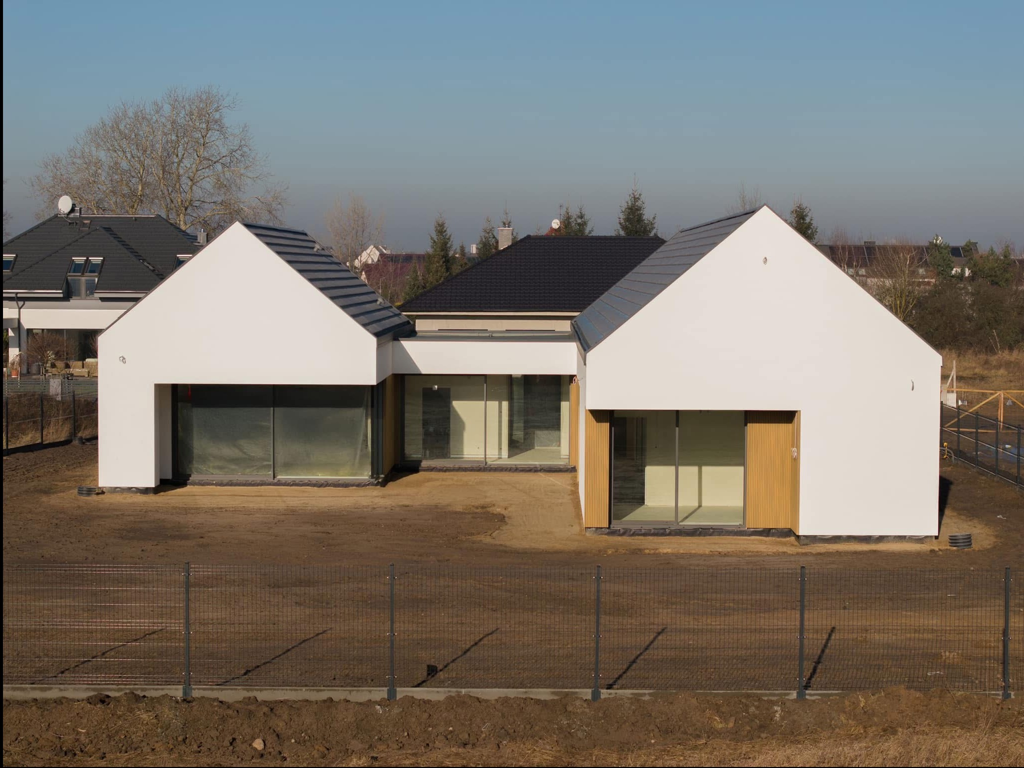 Nowoczesny parterowy dom z białymi ścianami i ciemną dachówką, otoczony ziemistym terenem i metalowym ogrodzeniem. Widoczne duże przeszklenia i minimalistyczny design.
