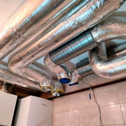 NOVUM-INSTAL - Instalacja wentylacyjna z izolowanymi przewodami i elementami metalowymi, zamontowana pod sufitem w pomieszczeniu z dwoma piecami grzewczymi.