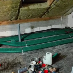 NOVUM-INSTAL - Montaż rekuperacji na poddaszu: zielone rury wentylacyjne ułożone na czarnej folii, izolacja wełną mineralną na skośnym stropie. Widoczne narzędzia i materiały instalacyjne.