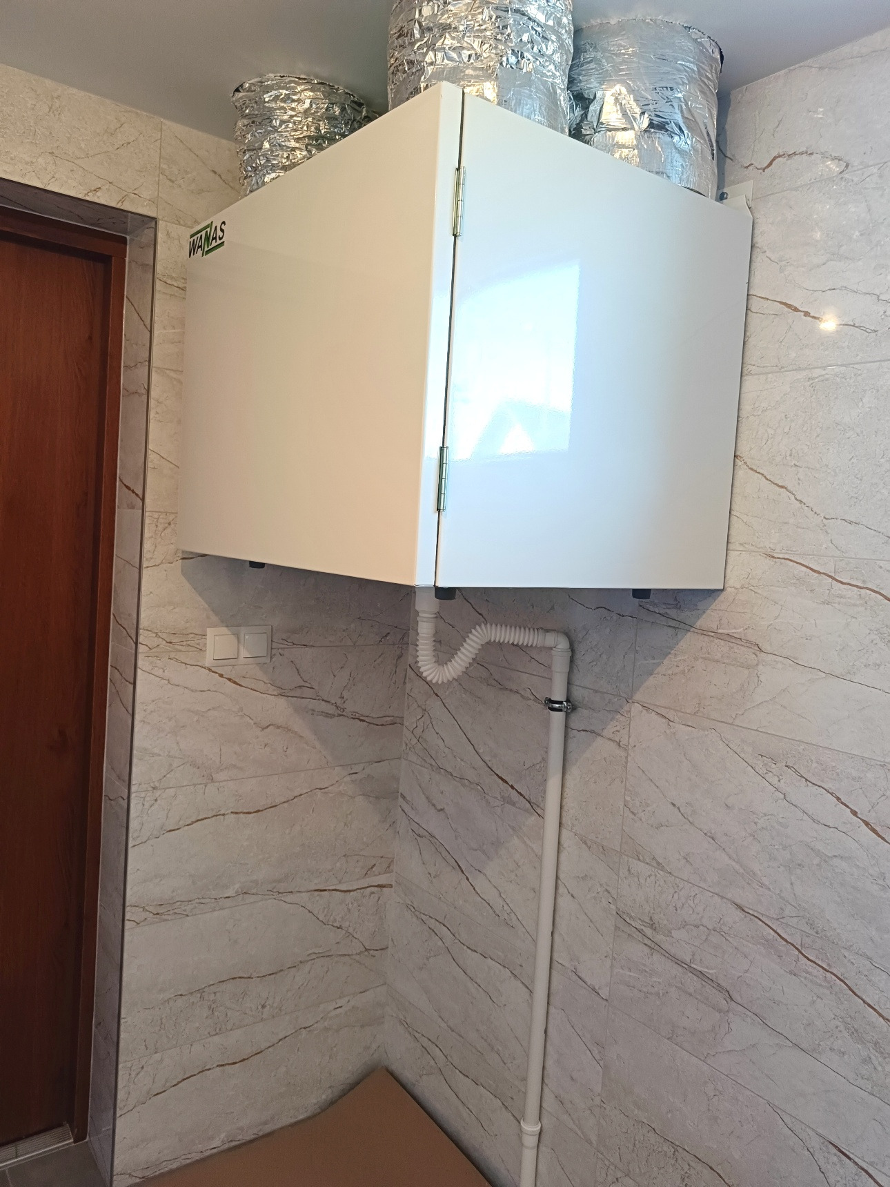 Biała jednostka rekuperacji marki Wanas zamontowana w rogu pomieszczenia z marmurowymi płytkami i widocznymi przewodami wentylacyjnymi.