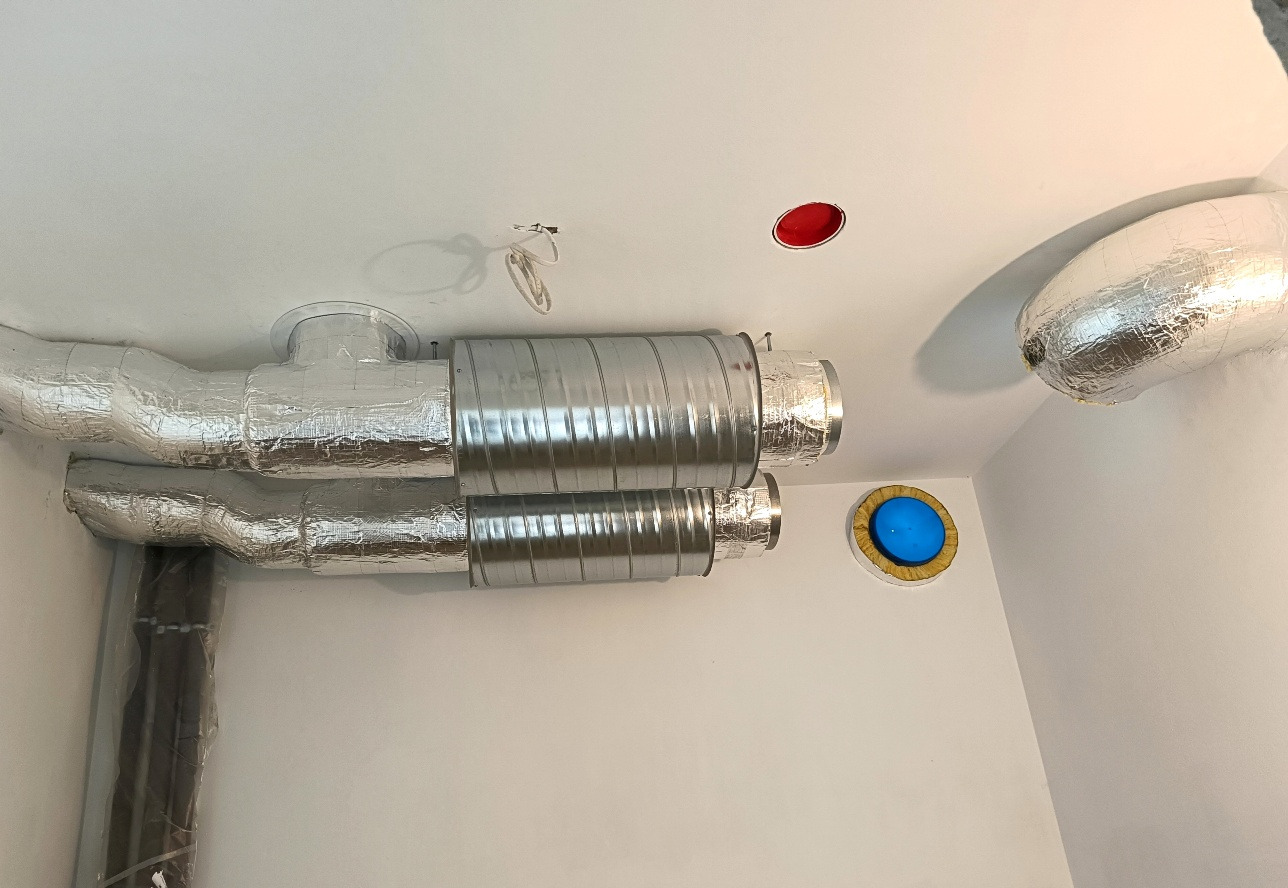Dwa równoległe przewody wentylacyjne z izolacją aluminiową, zamontowane pod sufitem. Widoczne otwory wentylacyjne i instalacja elektryczna.