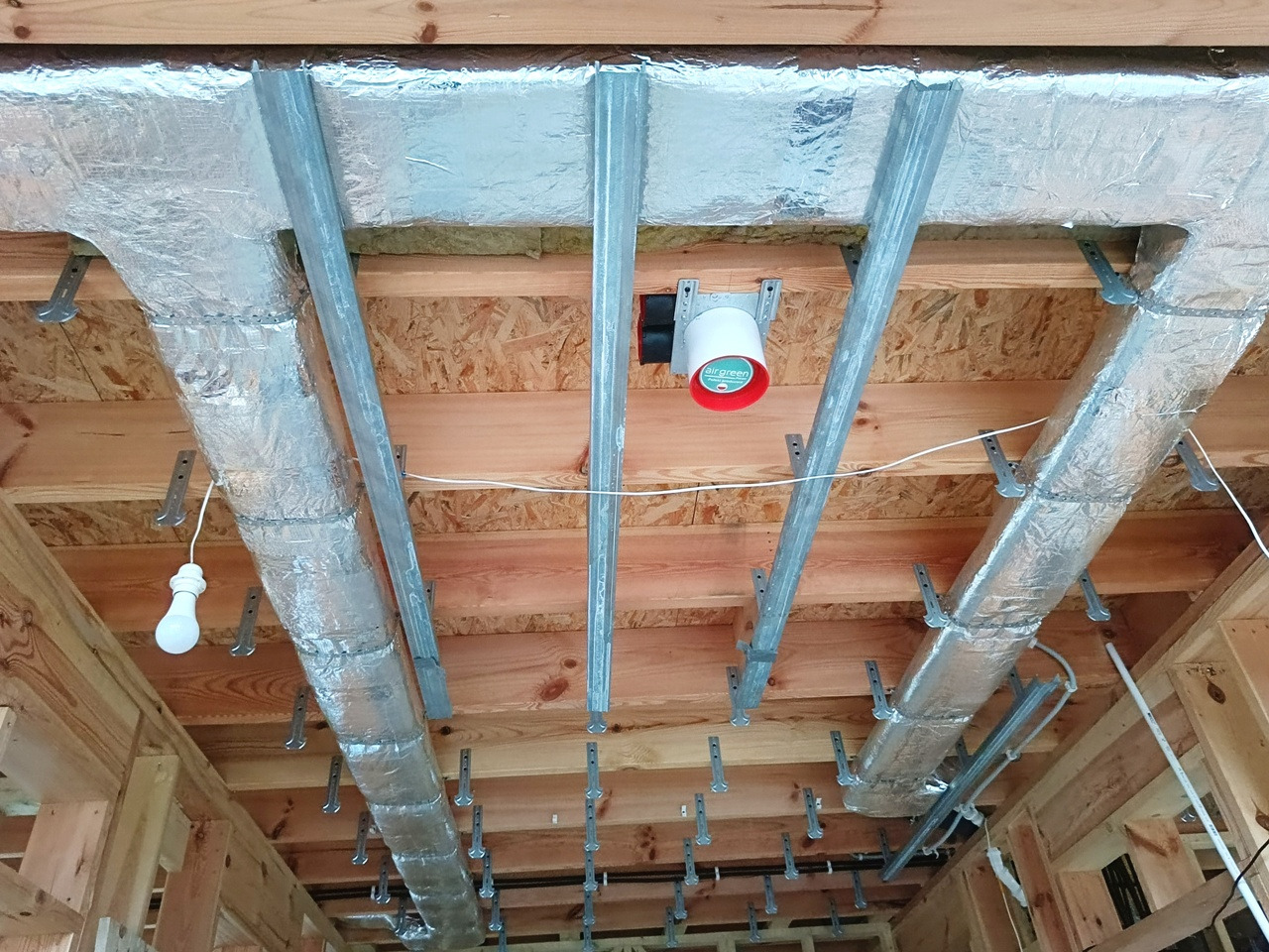 Montaż kanałów wentylacyjnych z izolacją aluminiową, widoczne metalowe profile mocujące i czujnik AirGreen. Instalacja pod stropem z drewnianych belek.