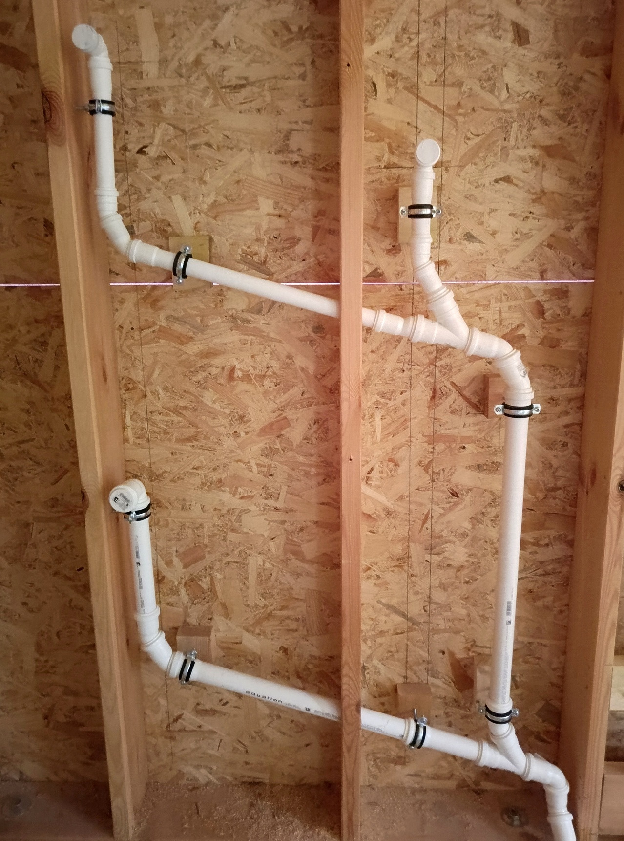 Białe rury wentylacyjne zamontowane w drewnianej konstrukcji ściany, zabezpieczone metalowymi obejmami. Widoczny laserowy poziom.