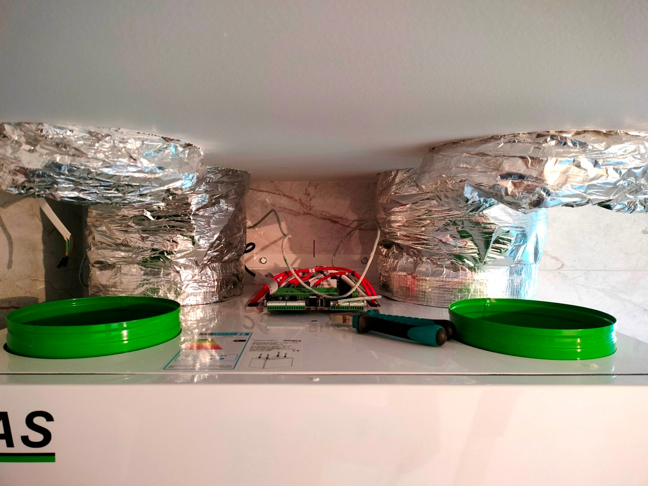 Wnętrze rekuperatora z podłączonymi przewodami wentylacyjnymi w folii aluminiowej. Widoczne elementy elektroniki i zielone pierścienie uszczelniające.