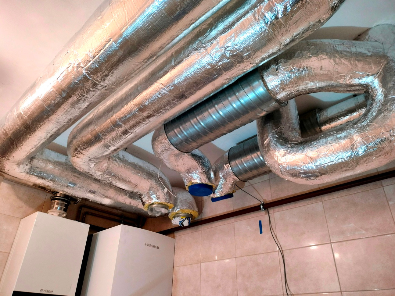 Instalacja wentylacyjna z izolowanymi przewodami i elementami metalowymi, zamontowana pod sufitem w pomieszczeniu z dwoma piecami grzewczymi.