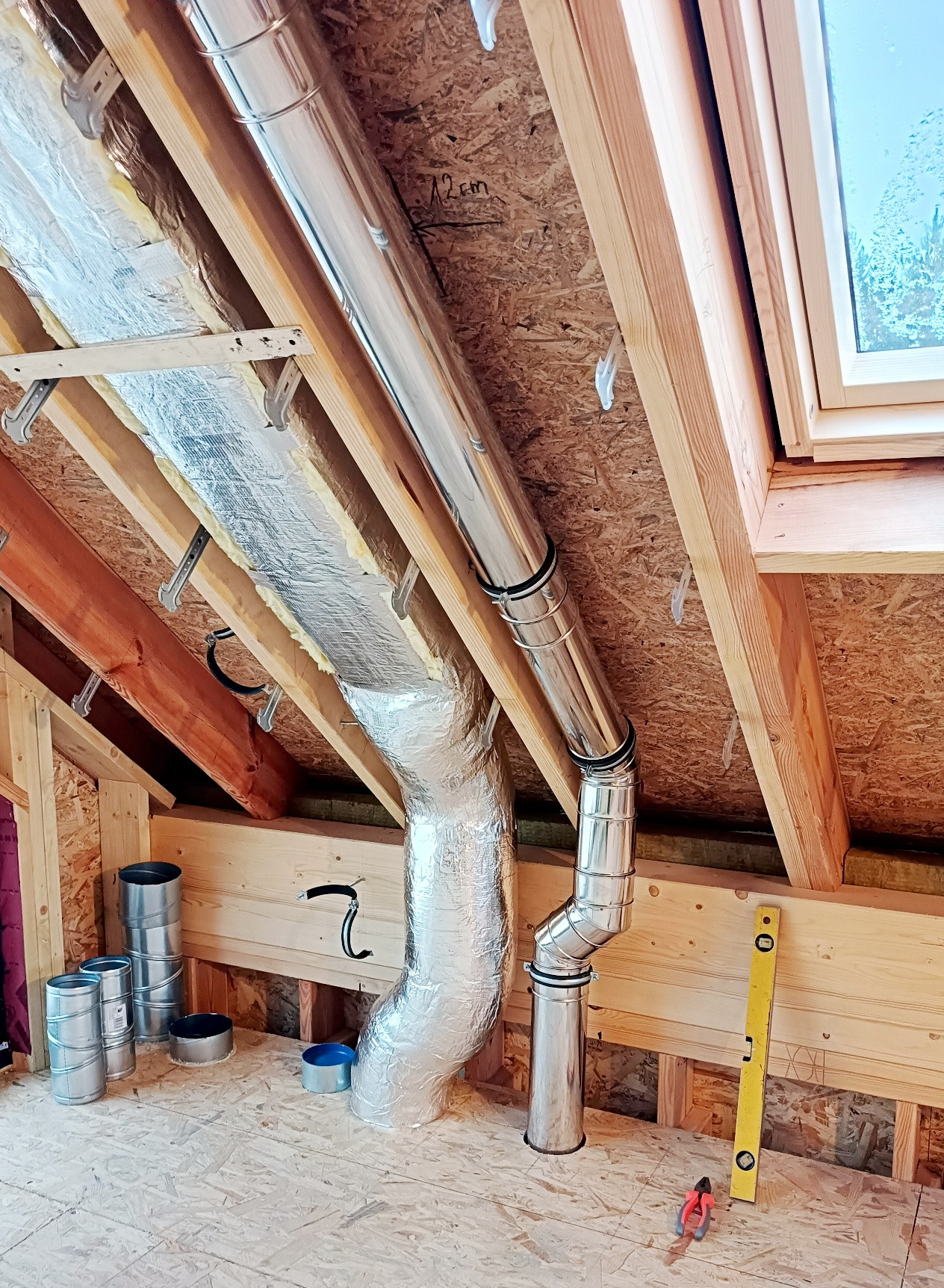 Montaż rur wentylacyjnych w surowym wnętrzu poddasza z oknem dachowym. Widoczne materiały izolacyjne i narzędzia. Elementy systemu wentylacji w trakcie instalacji.