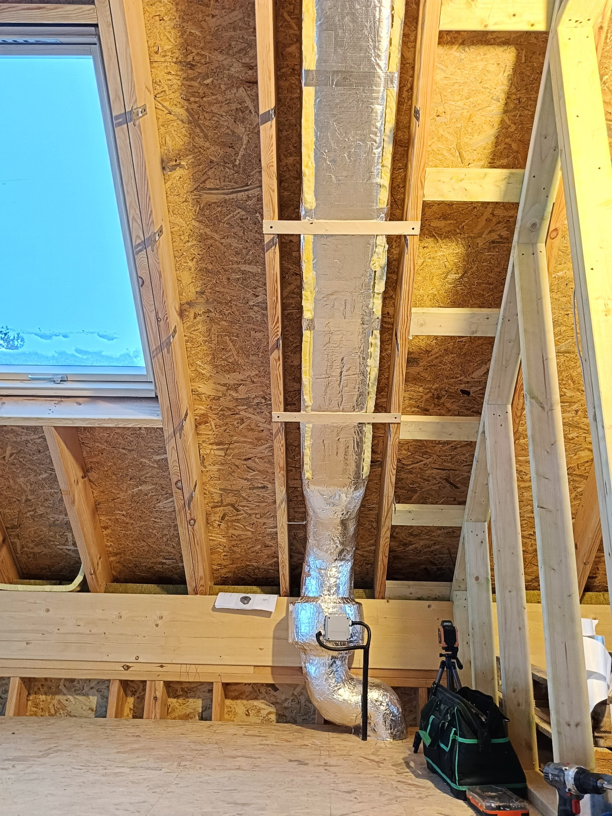 Montaż kanału wentylacyjnego w nowym budynku z oknem dachowym. Widoczne drewniane elementy konstrukcyjne i narzędzia. Izolowany kanał wentylacyjny biegnie pionowo.