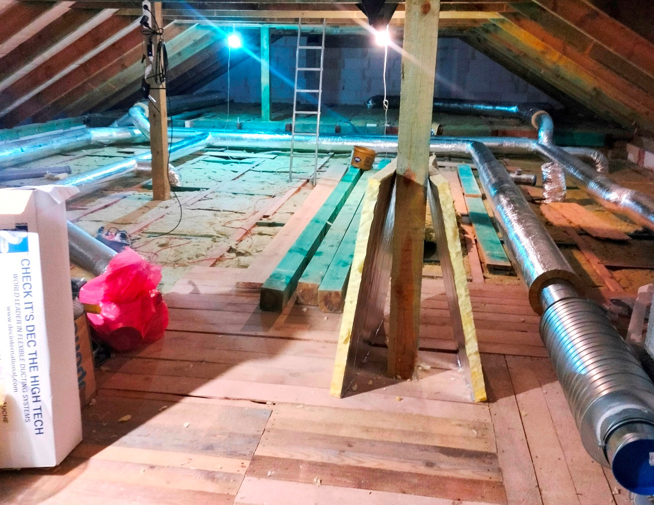 Montaż instalacji wentylacyjnej na poddaszu. Widoczne izolowane rury wentylacyjne, drewniane elementy konstrukcyjne i materiały izolacyjne. Etap prac w trakcie realizacji.