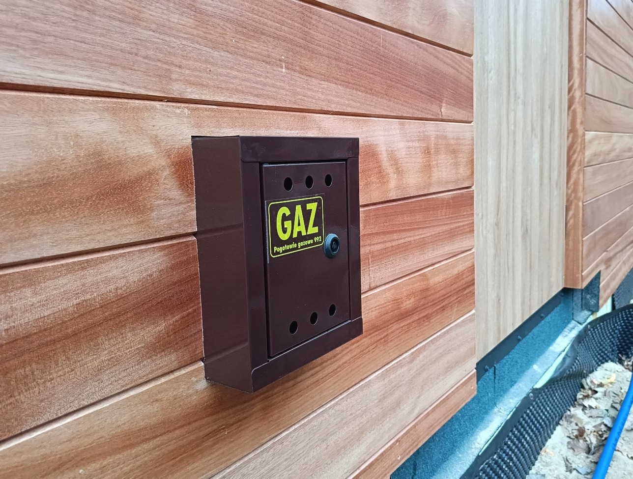 Brązowa skrzynka gazowa z żółtą naklejką 'GAZ' zamontowana na elewacji z drewnianych paneli. Widoczne elementy instalacji przy gruncie.