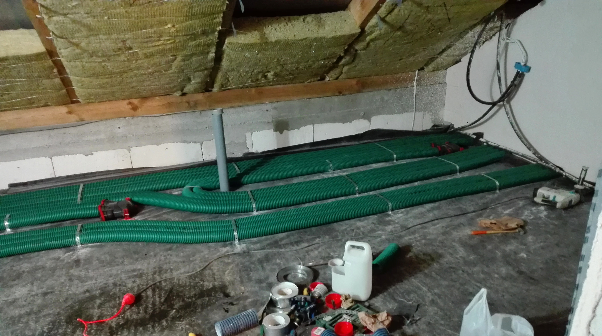 Montaż rekuperacji na poddaszu: zielone rury wentylacyjne ułożone na czarnej folii, izolacja wełną mineralną na skośnym stropie. Widoczne narzędzia i materiały instalacyjne.