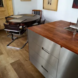 Nowoczesna kuchnia: drewniany blat z metalowymi szafkami, okrągły stół z krzesłami, drewniana podłoga i grafiki na ścianach tworzą przytulne wnętrze.