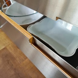 Witold Szostak - Szostak Atelier - Otwarta szuflada z metalowym frontem i drewnianymi bokami, wypełniona naczyniami. Widoczny fragment drewnianej podłogi i blat.