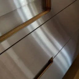 Metalowe fronty szuflad z subtelnymi, drewnianymi uchwytami wpuszczonymi w powierzchnię, ukazane pod kątem, z widocznymi refleksami światła.