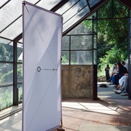 Witold Szostak - Szostak Atelier - Parawan z białym płótnem w szklarni, wsparty na drewnianych nóżkach. W tle siedzący ludzie i zieleń. Geometryczny wzór z cienkich linek na płótnie.