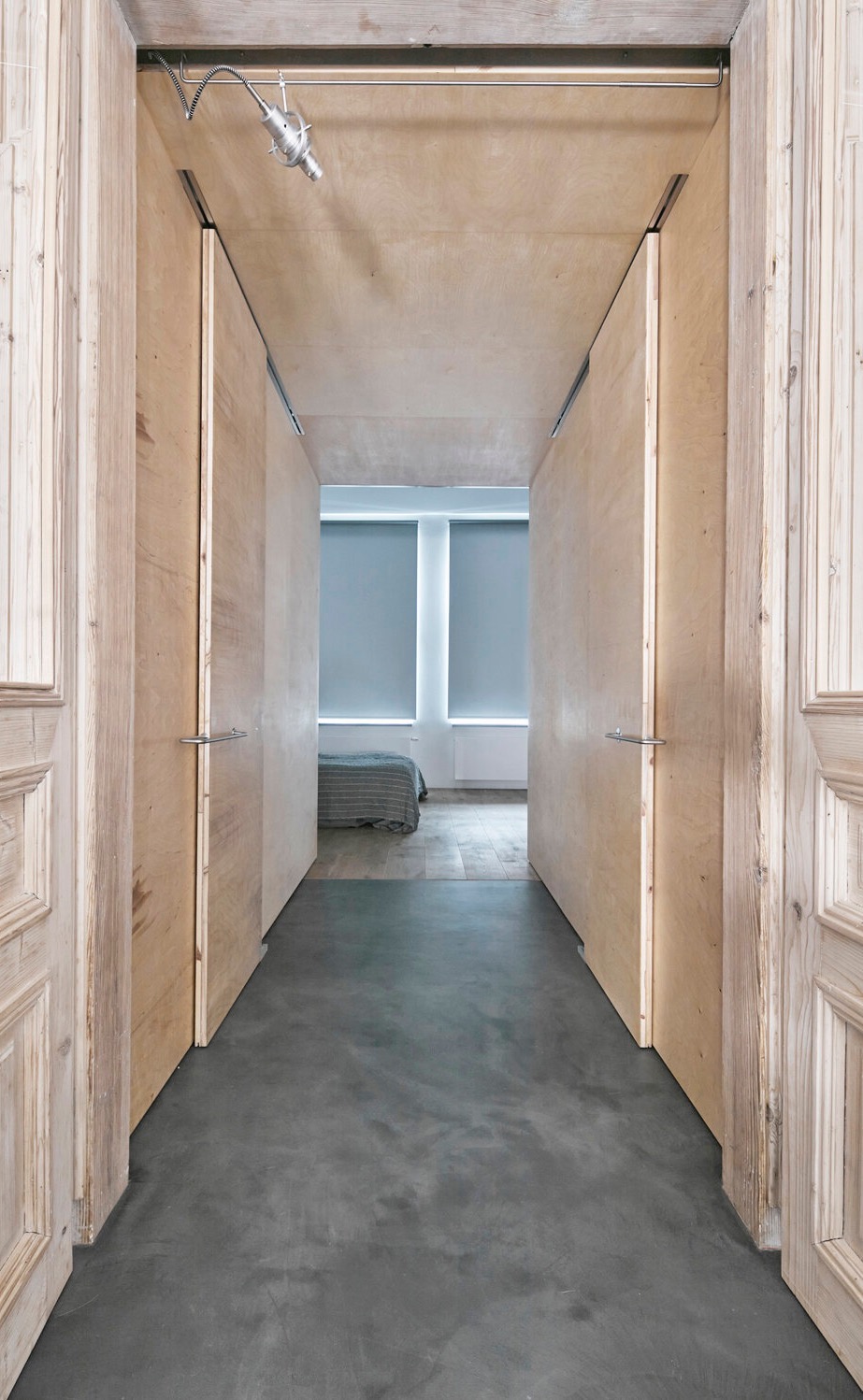 Wnętrze z otwartymi, przesuwnymi drzwiami z surowego drewna, ukazujące widok na sypialnię z szarymi roletami i betonową podłogą. Minimalistyczny design i surowe wykończenie.