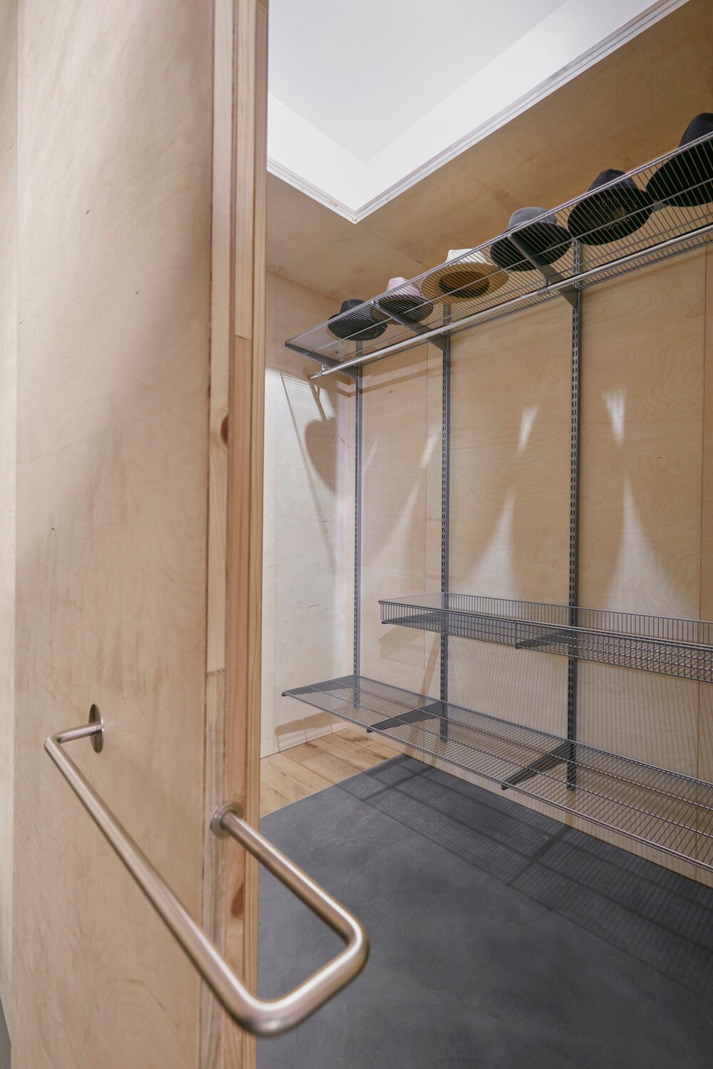 Otwarta, jasna garderoba z metalowymi półkami i drzwiami z uchwytem. Na górnej półce ułożone kapelusze. Ściany wyłożone sklejką. Minimalistyczny design.