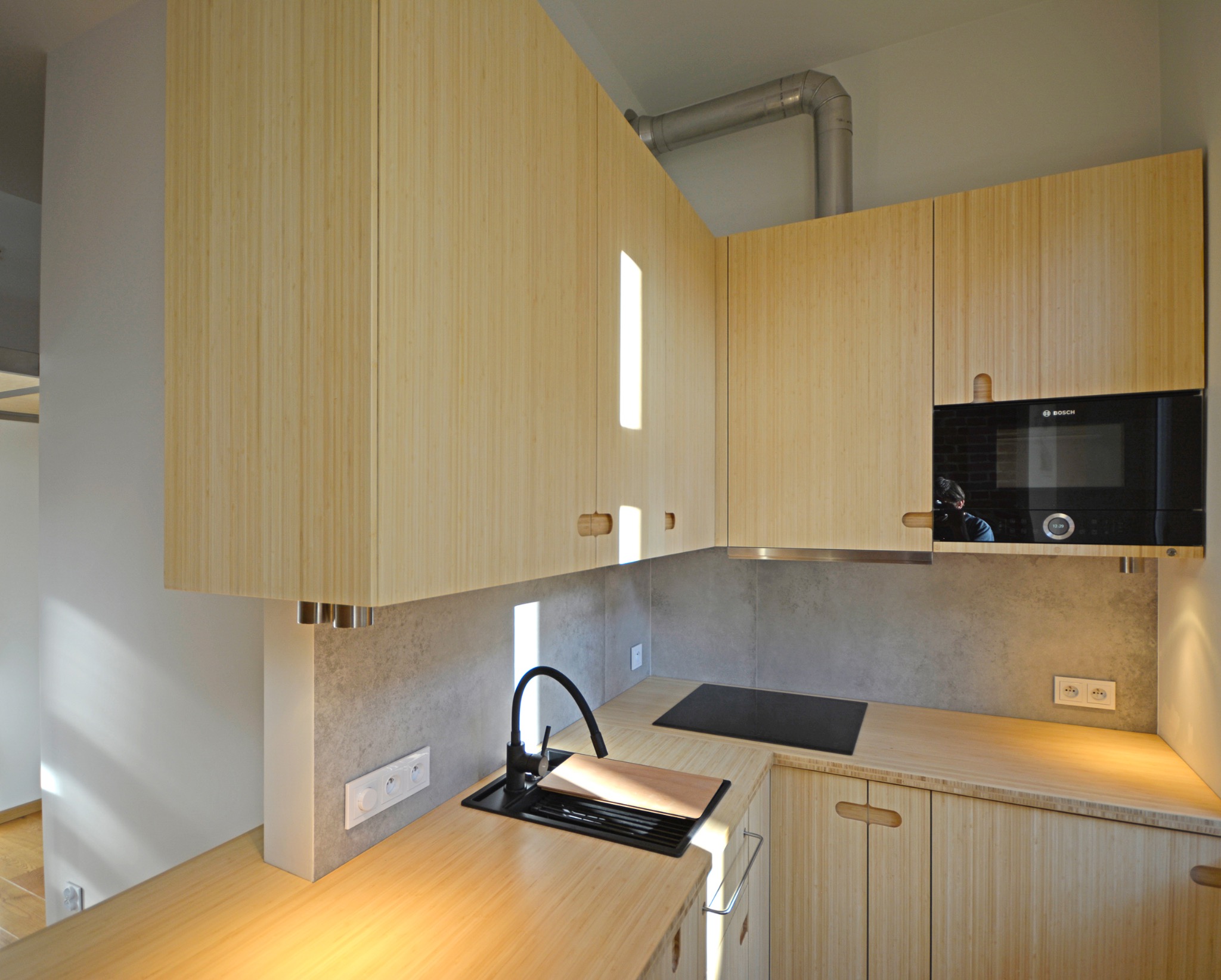 Jasna kuchnia z bambusowymi frontami szafek, czarnym zlewem i płytą grzewczą. Widoczne gniazdka elektryczne i mikrofalówka Bosch. Minimalistyczny design z drewnianymi uchwytami.