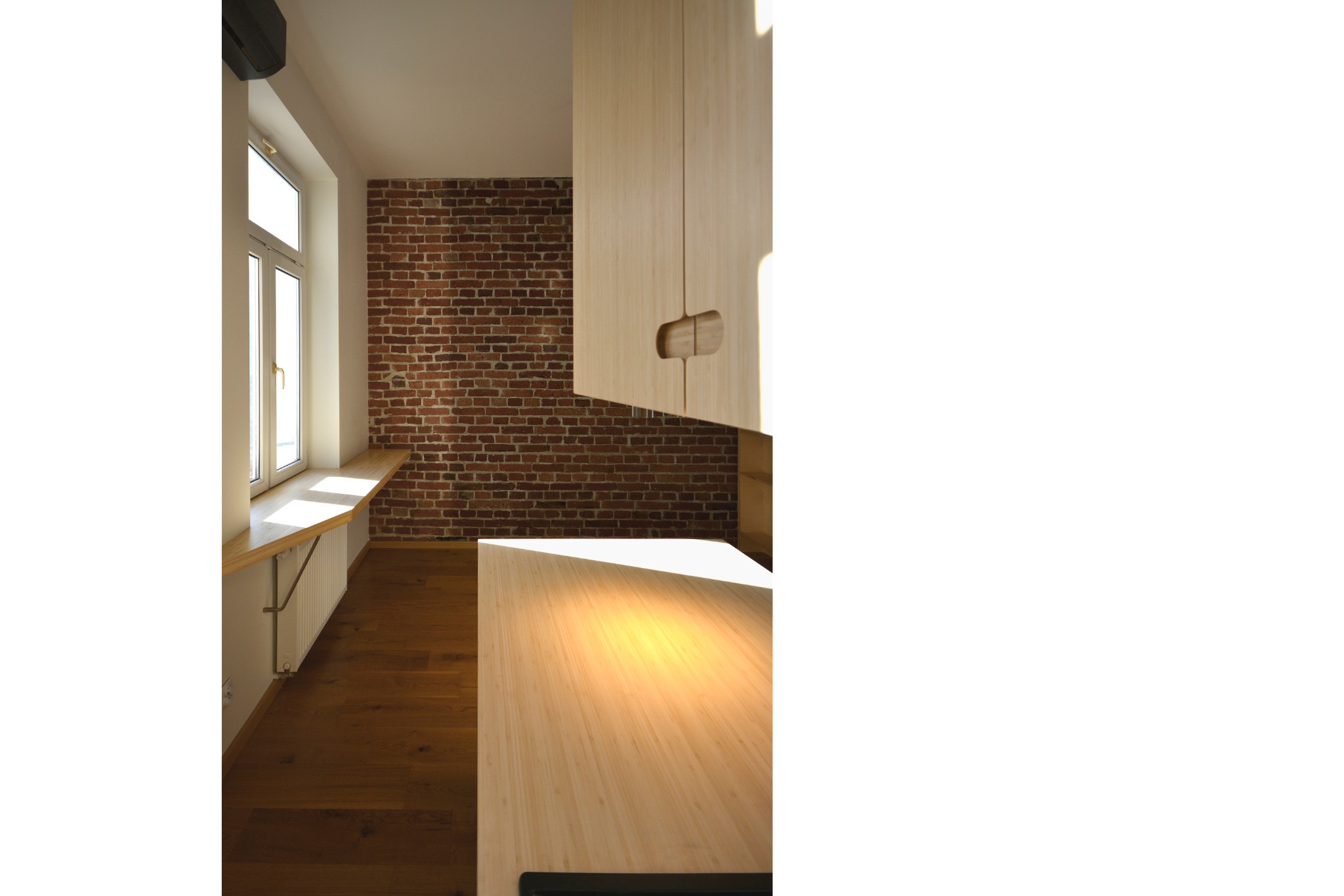 Jasna kuchnia z drewnianym blatem i szafkami, ceglana ściana w tle. Parapet drewniany pod oknem. Minimalistyczny design, ciepłe oświetlenie.