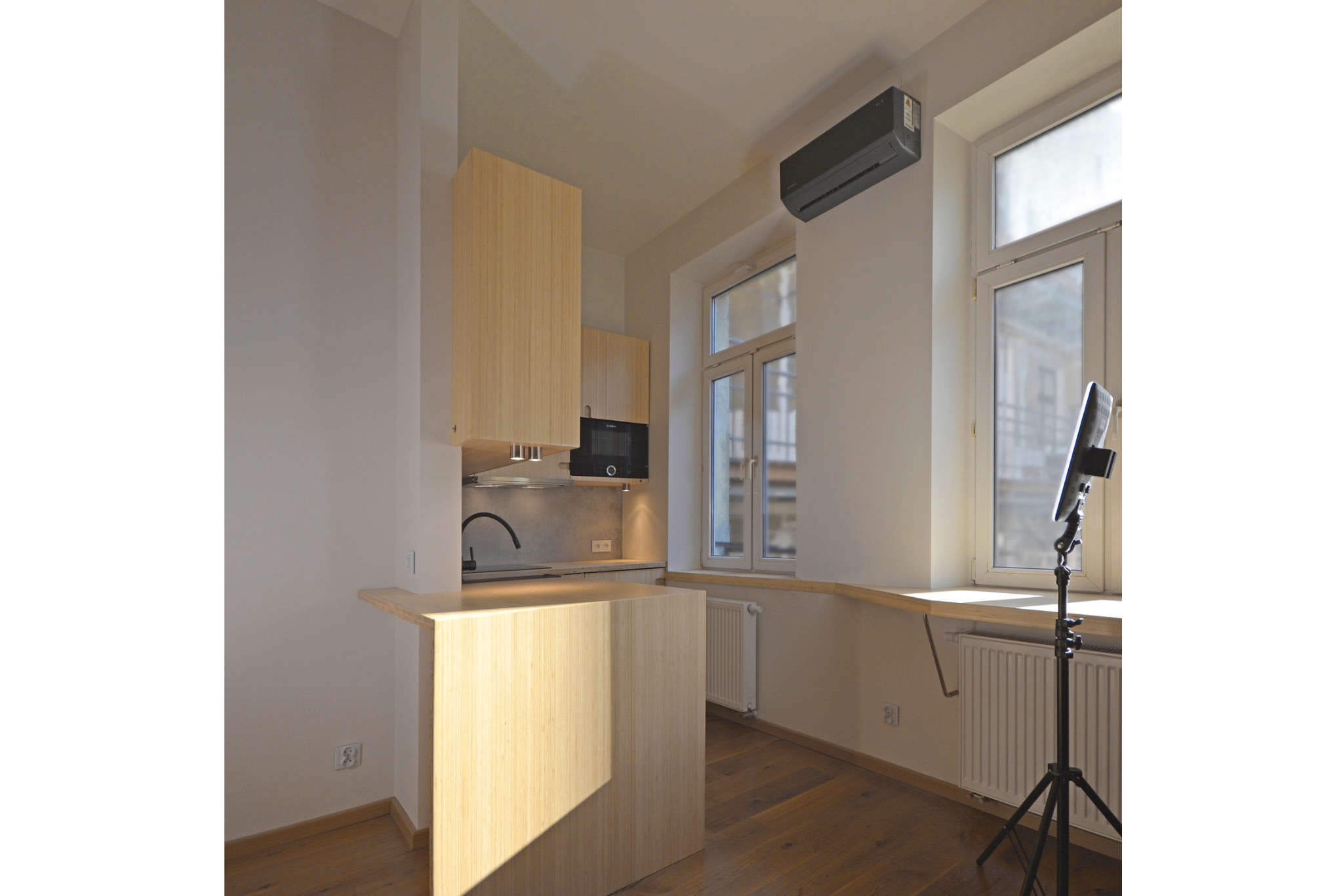Jasna kuchnia z drewnianymi frontami, wyspą i sprzętem AGD. Uwagę zwraca lampa studyjna stojąca przy oknie, sugerująca profesjonalną sesję zdjęciową wnętrza.