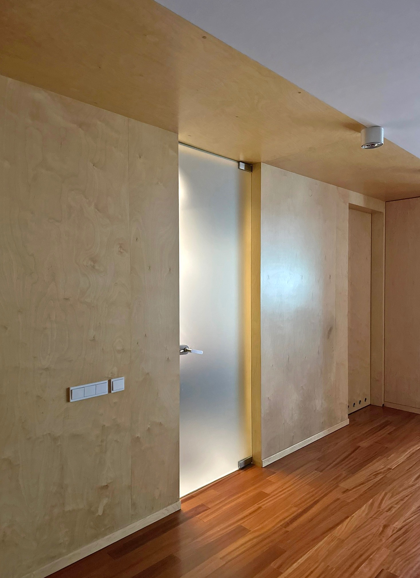 Wnętrze domu wykończone sklejką: ściany, sufit i drzwi przesuwne. Widoczny fragment drewnianej podłogi i włączniki światła. Minimalistyczny design z naturalnych materiałów.