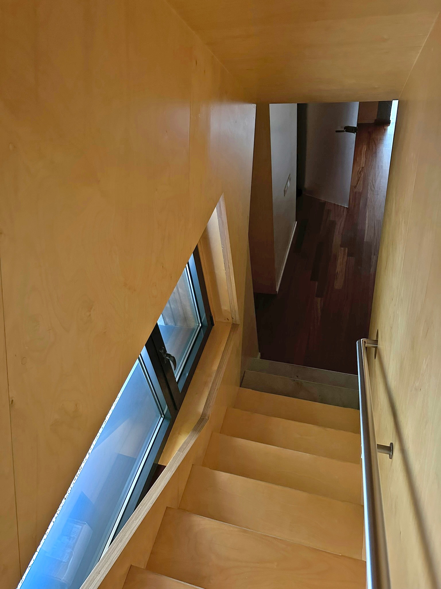 Klatka schodowa wykończona drewnem z oknem o nietypowym kształcie, widok z góry. Elementy ze stali nierdzewnej. W tle widoczne wejście do pomieszczenia z drewnianą podłogą.