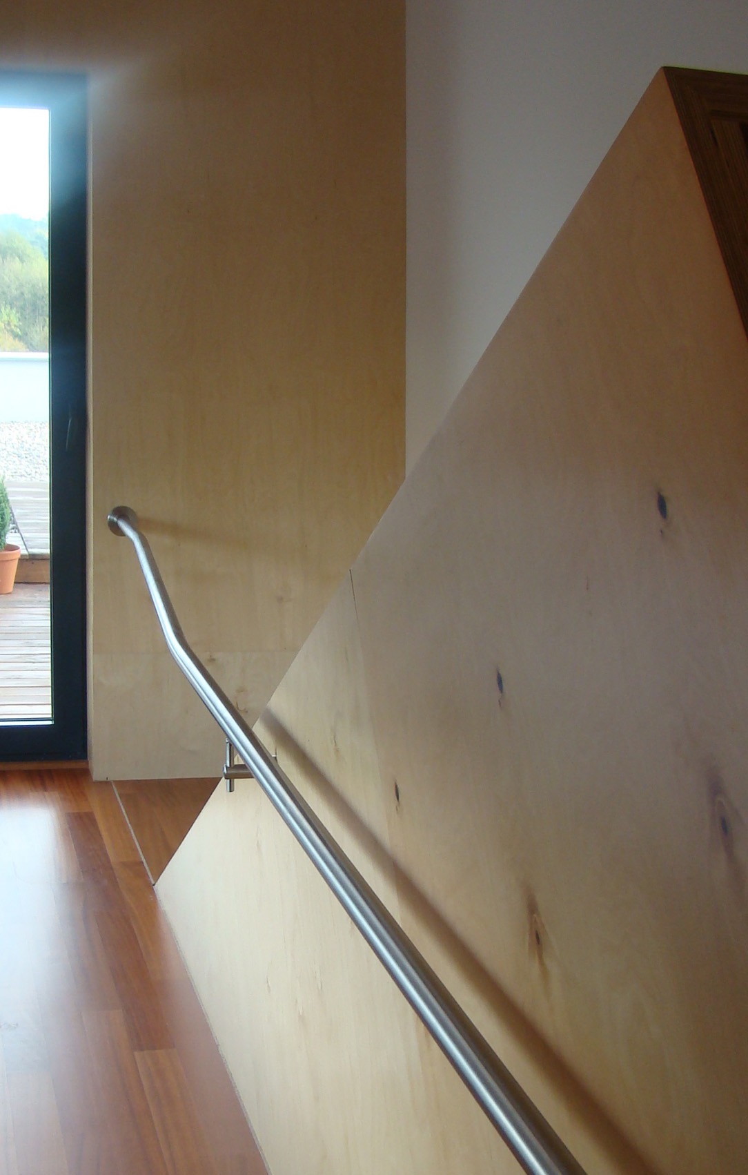 Nowoczesne schody z drewnianą zabudową i metalową poręczą, widok z dołu, z bliska. Na podłodze parkiet. Za oknem zieleń.