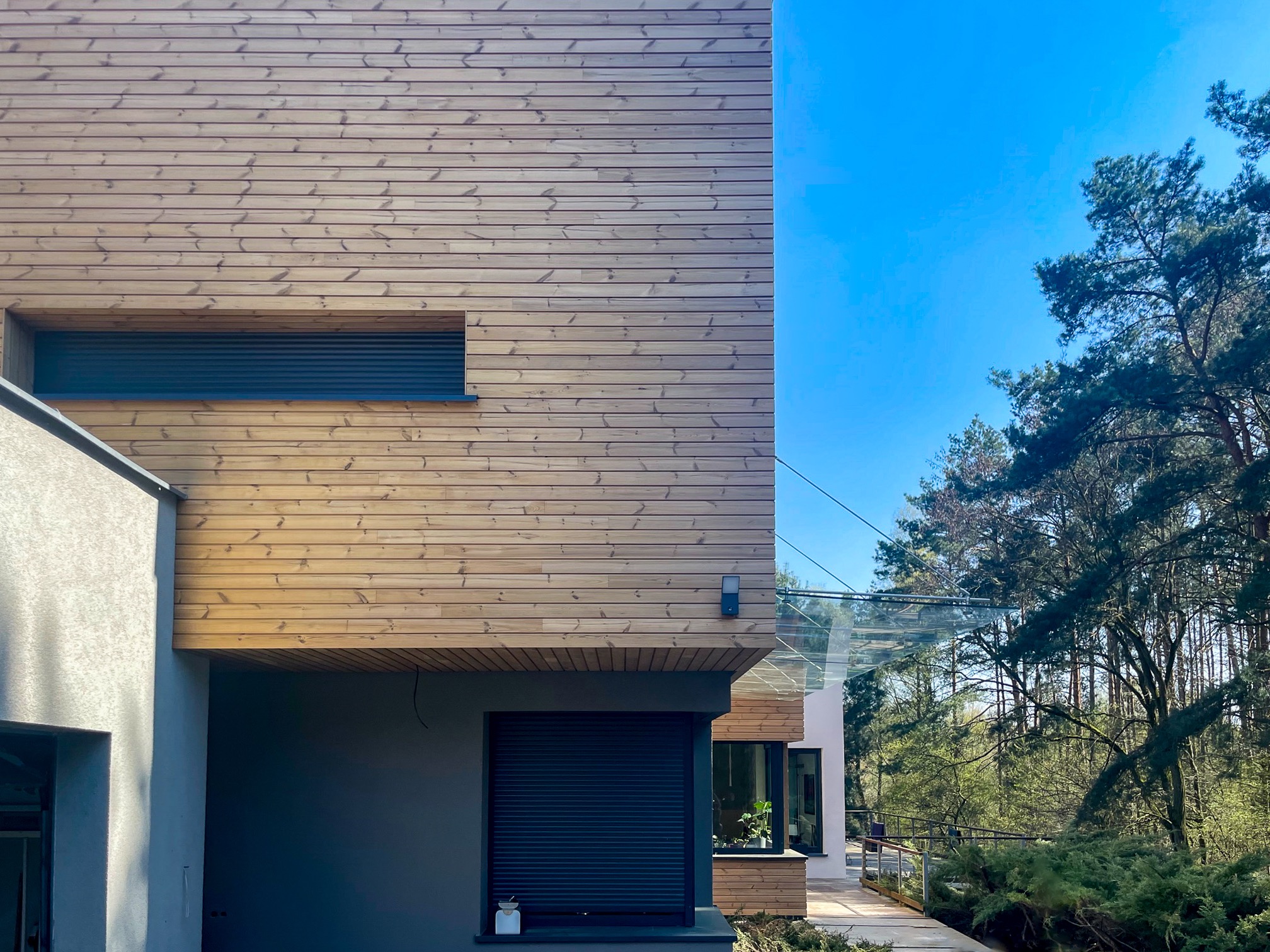 Elewacja nowoczesnego domu z drewnianą okładziną i roletami zewnętrznymi, na tle lasu i błękitnego nieba. Widoczne detale architektoniczne i naturalne otoczenie.