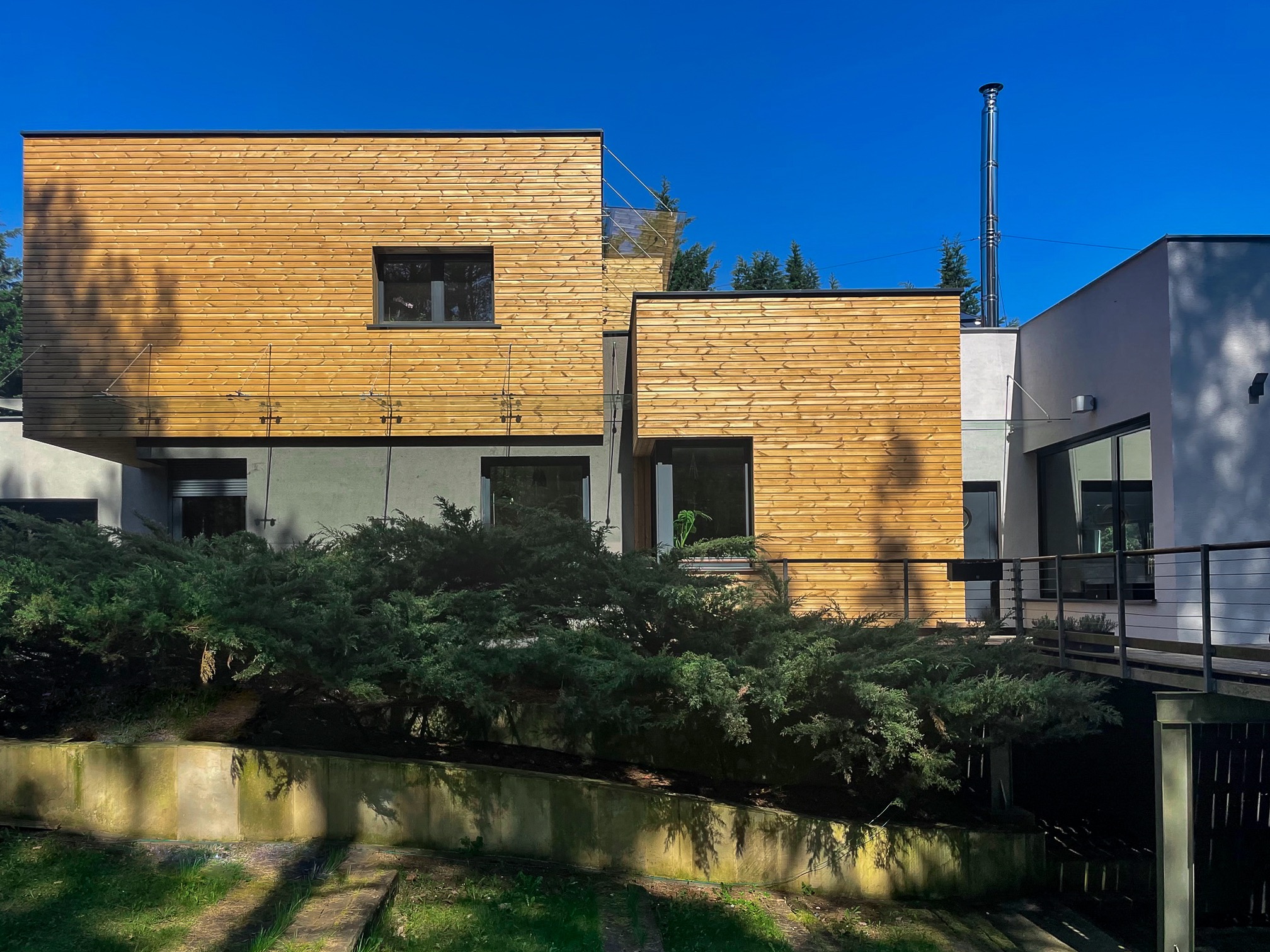 Nowoczesna willa z drewnianą elewacją i dużymi oknami, otoczona zielenią. Widoczny podjazd i fragment mostku.