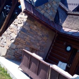 DW-KAM Daniel Wójcik - Kamienna elewacja domu z tarasem i widocznym fragmentem dachu zabezpieczonego folią. Na tarasie stoi brązowa sofa.