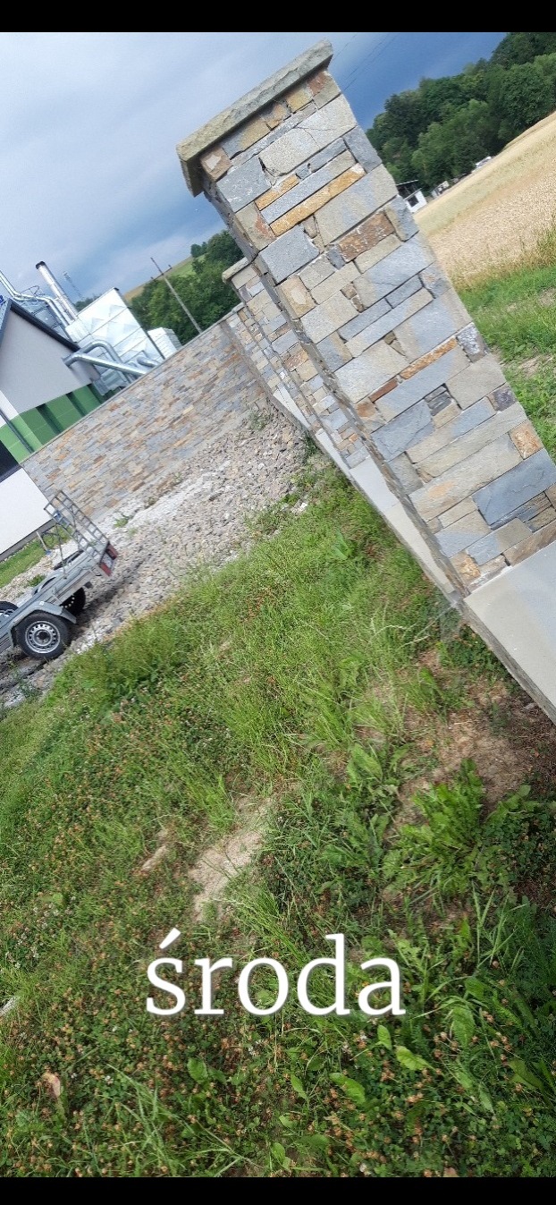 Kamienne słupki ogrodzeniowe z nieregularnych płytek, widoczne z perspektywy trawnika. W tle budynek i pole.