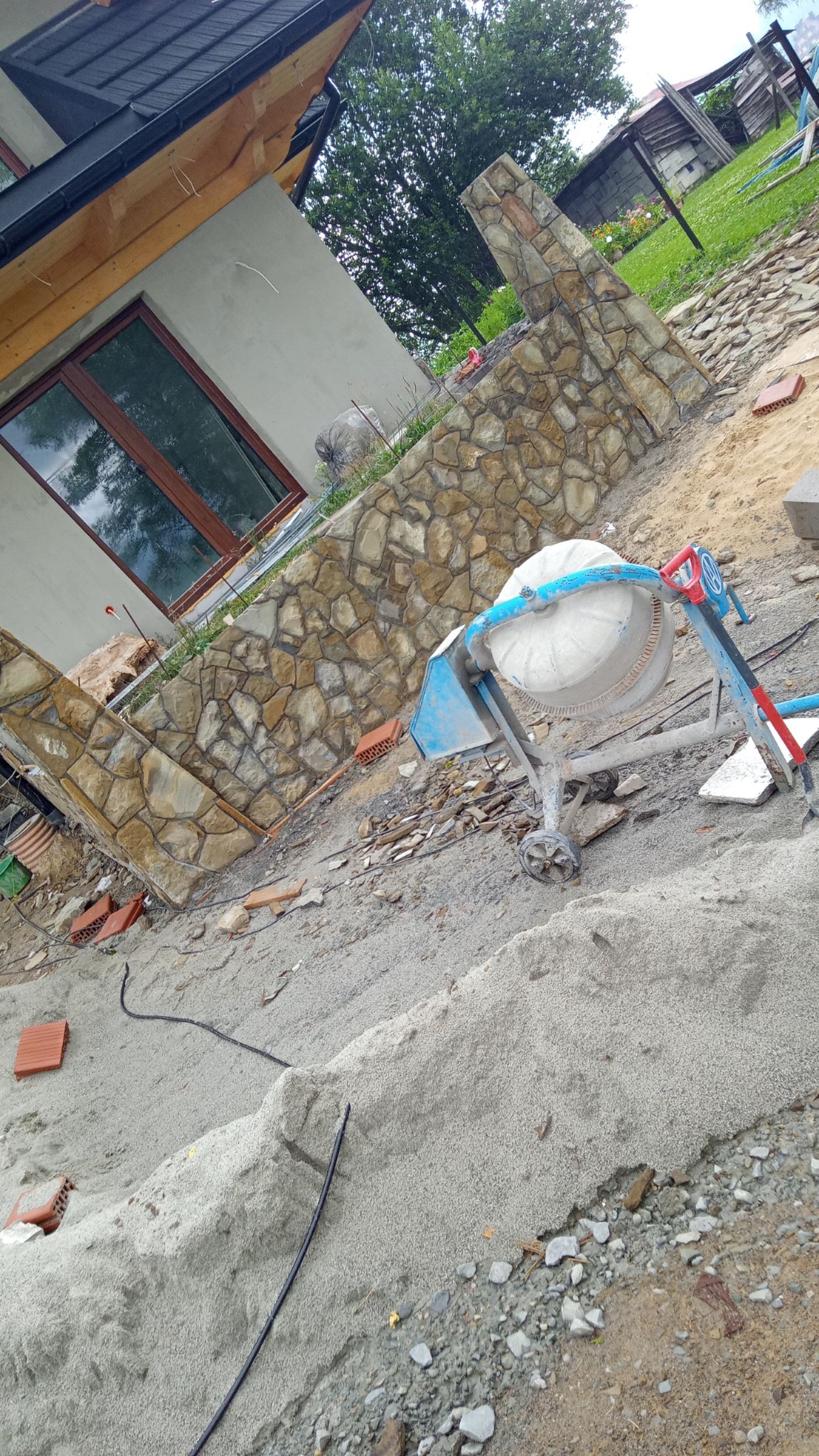 Budowa muru z kamienia łupanego przy domu. Widoczna betoniarka i materiały budowlane. Częściowo ukończony fragment elewacji z kamienia.