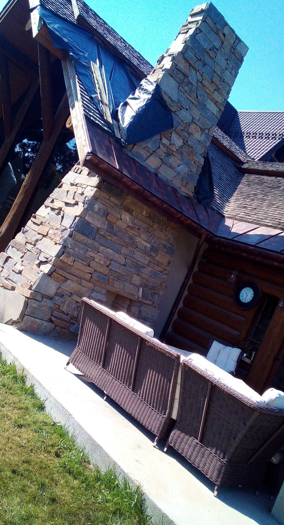 Kamienna elewacja domu z tarasem i widocznym fragmentem dachu zabezpieczonego folią. Na tarasie stoi brązowa sofa.