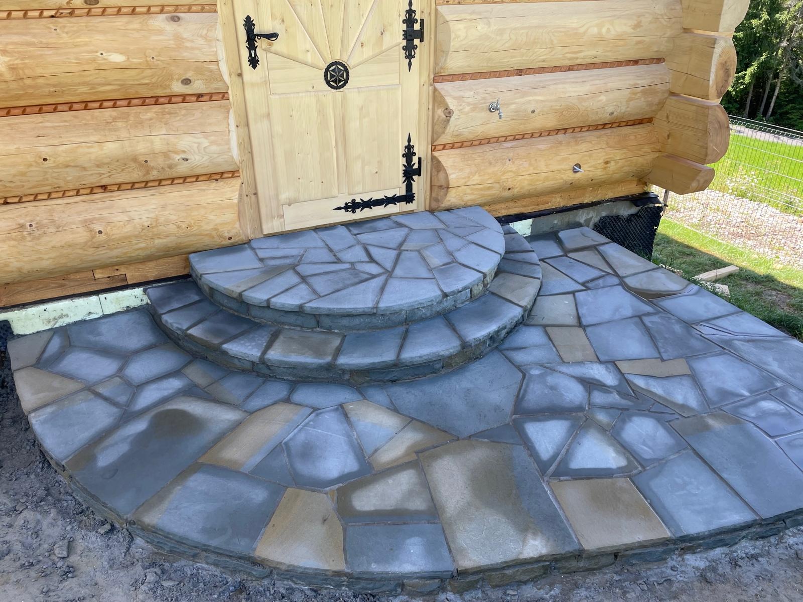 Kamienne schody wejściowe, wykonane z nieregularnych płyt, prowadzą do drewnianego domu z dekoracyjnymi okuciami drzwi. Widok z dołu, podkreślający fakturę kamienia.