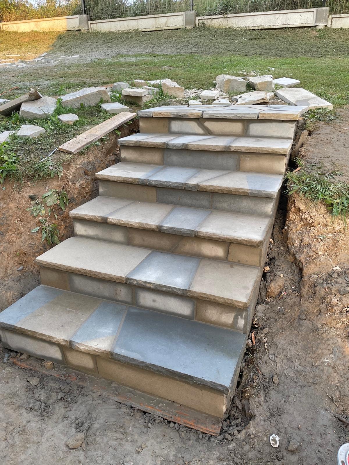 Kamienne schody zewnętrzne w trakcie budowy, widoczne niedokończone obrzeża i zapas materiału. Ujęcie z dołu, podkreślające solidność konstrukcji.