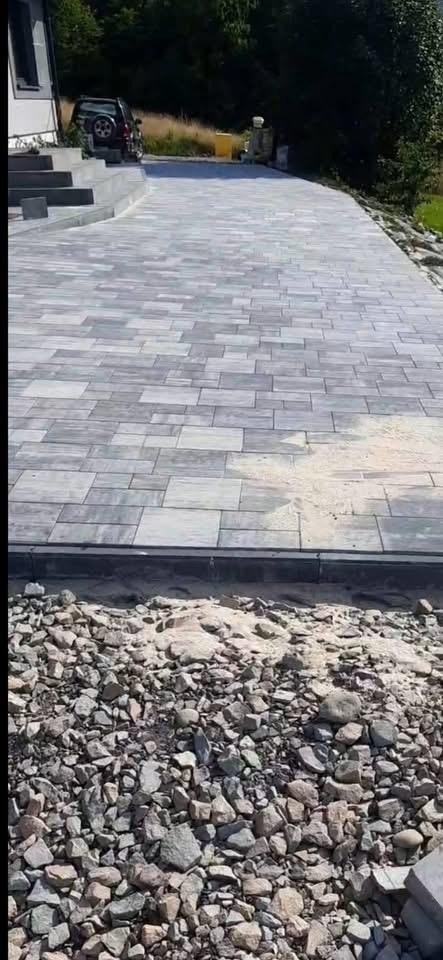 Szeroki podjazd z szarej kostki brukowej ułożonej w regularny wzór. W tle widoczne schody i częściowo zaparkowany samochód. Na pierwszym planie kamienie i piasek.