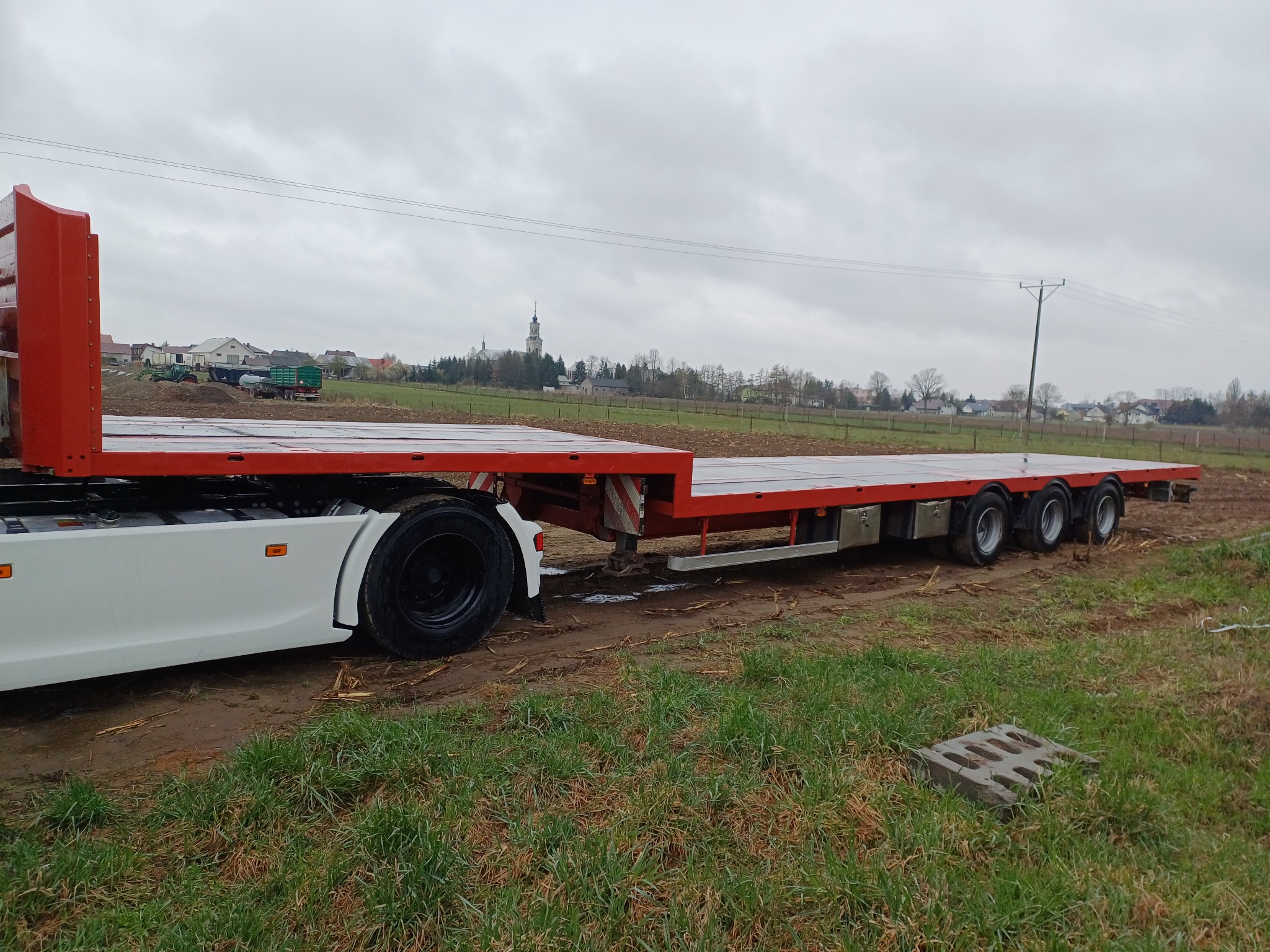 Czerwona naczepa niskopodwoziowa na polnej drodze, widok z boku na tle wiejskiego krajobrazu pod pochmurnym niebem. Widoczne koła i elementy konstrukcyjne.