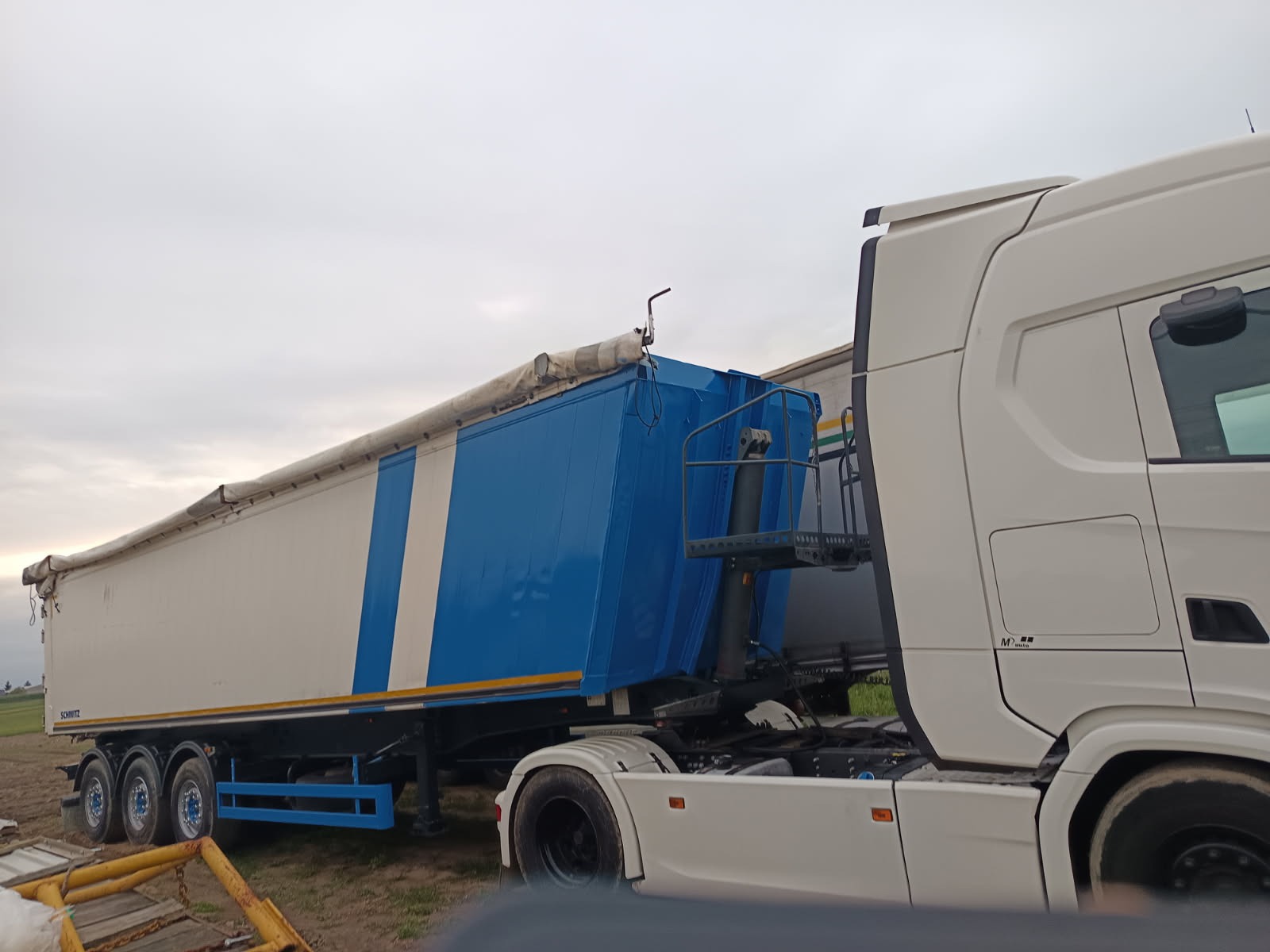 Ciężarówka z naczepą typu wywrotka, biało-niebieska, z widocznym podwoziem i oponami, zaparkowana na polu pod pochmurnym niebem.
