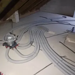 Szare, elastyczne rury wentylacyjne ułożone na poddaszu, prowadzące do rozdzielacza. Widoczne drewniane elementy konstrukcji dachu i betonowa posadzka.