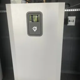 Biała pompa ciepła Thermia w kotłowni z szarą, perforowaną ścianą w tle, widoczne rury instalacyjne i skrzynka elektryczna. Ujęcie z dołu.