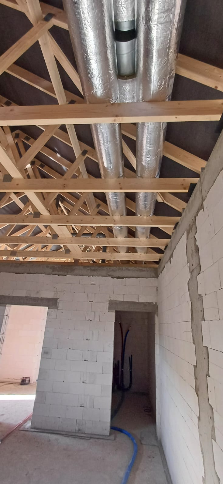 Montaż instalacji wentylacyjnej z izolowanymi rurami w surowym wnętrzu budynku z drewnianą konstrukcją dachu i ścianami z białych bloczków.