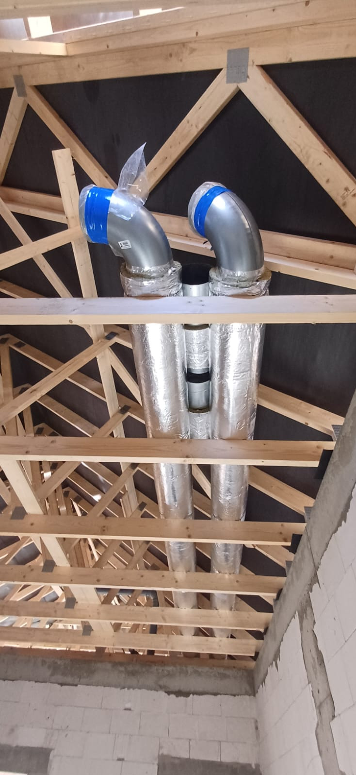 Dwie rury wentylacyjne z niebieską taśmą, owinięte folią, zamontowane pod drewnianą konstrukcją dachu w surowym wnętrzu budynku.