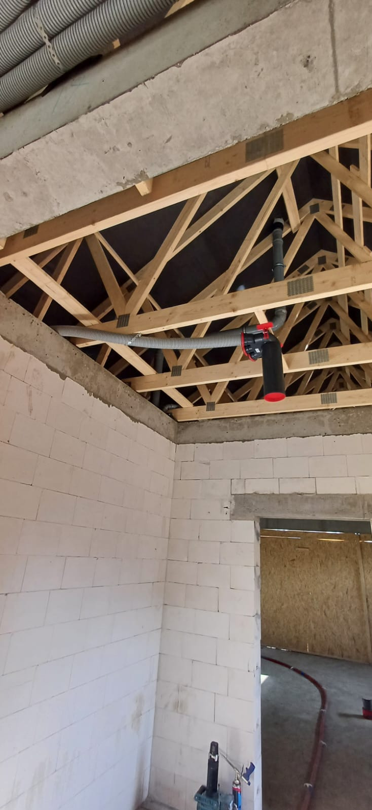 Szara rura wentylacyjna pod drewnianą konstrukcją dachu w surowym wnętrzu budynku z białego pustaka. Element instalacji rekuperacji z czarnym filtrem.