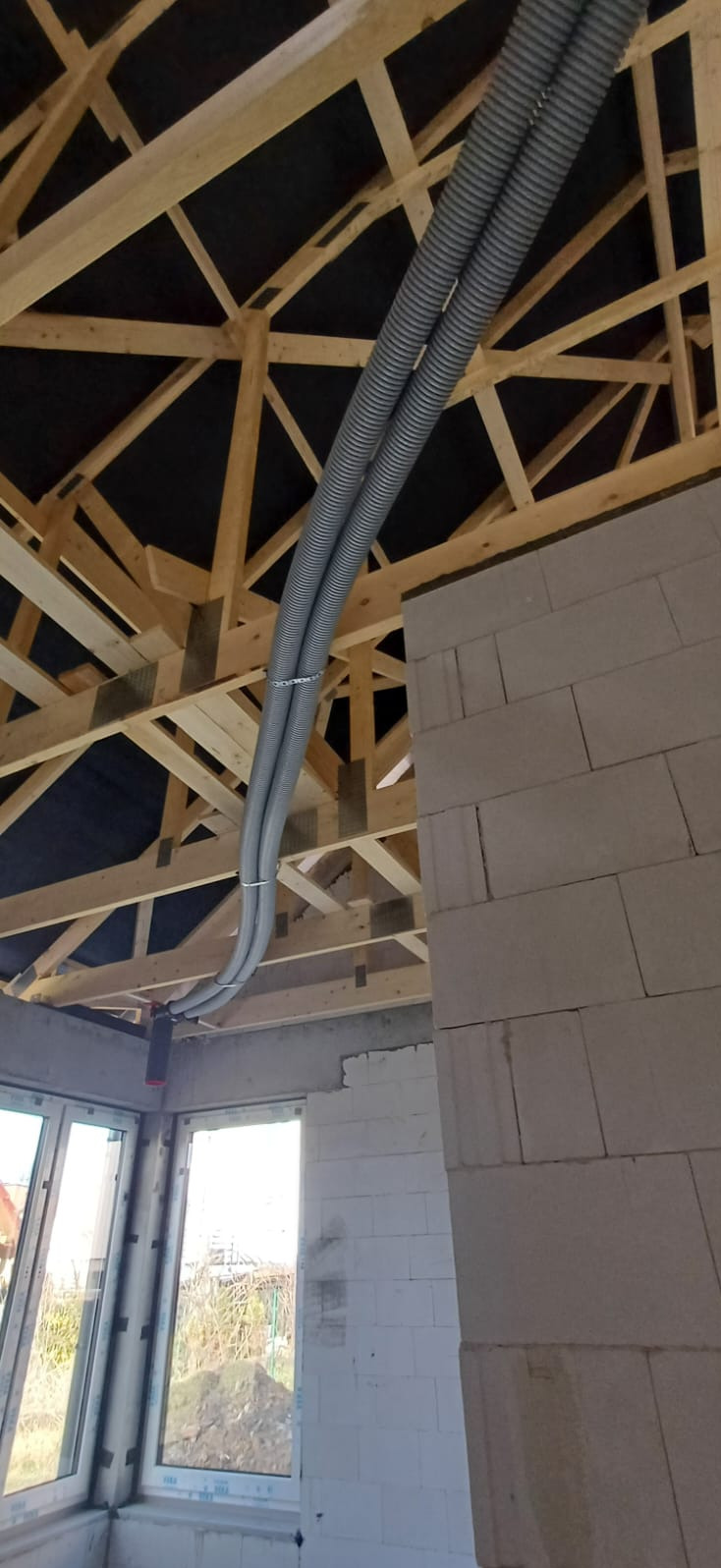 Dwie szare, karbowane rury przymocowane do drewnianej konstrukcji dachu w surowym stanie. Widoczne ściany z białych bloczków i okna. Instalacja w trakcie budowy.