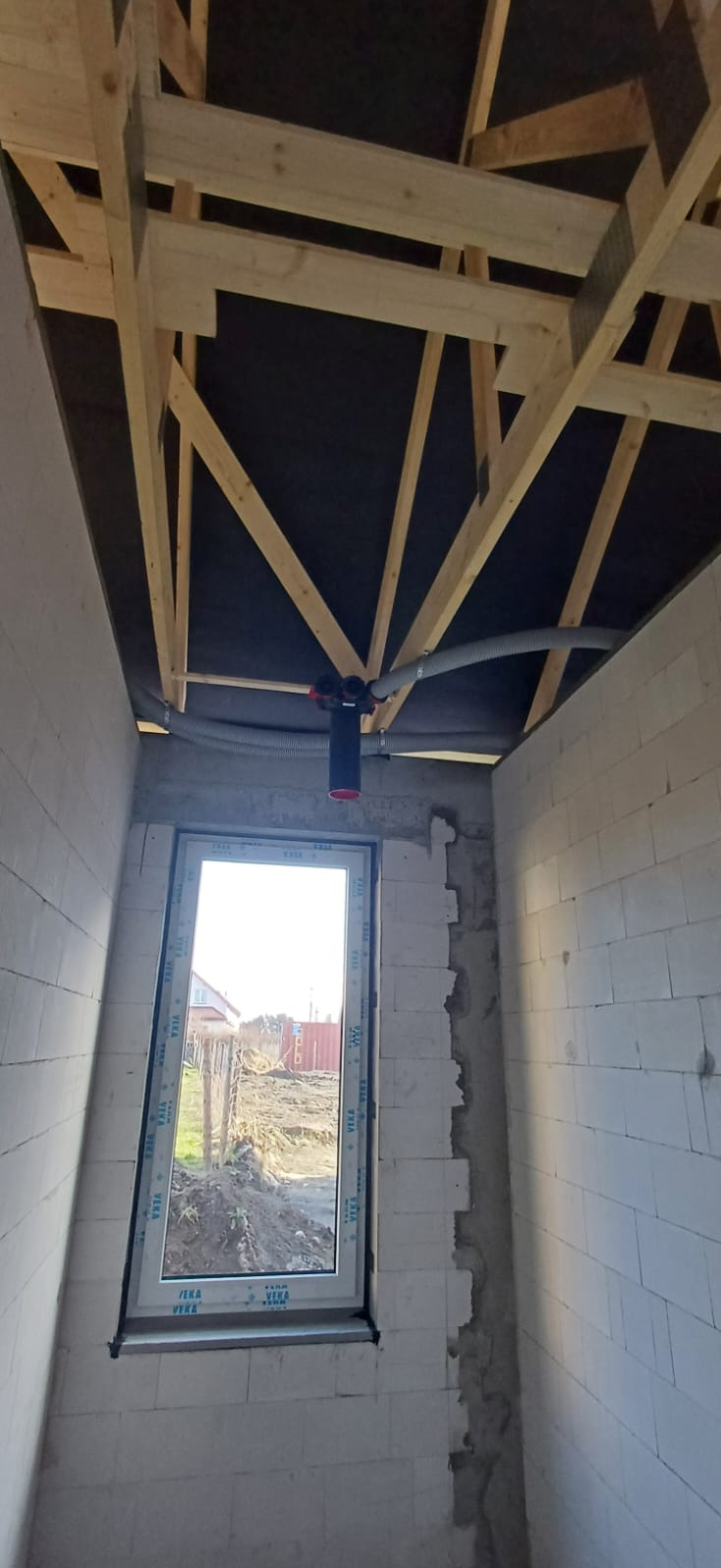 Wnętrze w trakcie budowy: widoczne drewniane elementy konstrukcji dachu, szara rura wentylacyjna i okno z widokiem na zewnątrz. Ściany z białych pustaków.