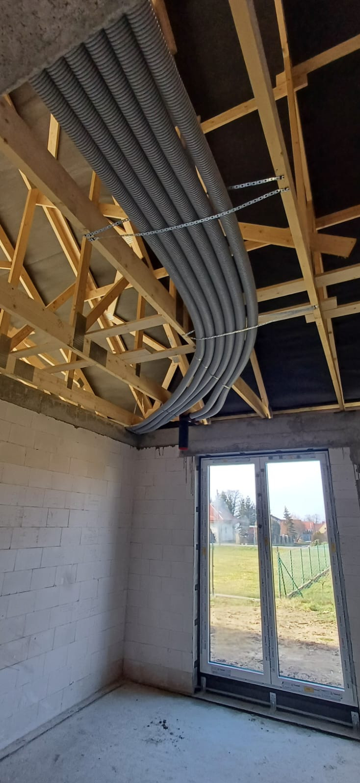 Szare rury wentylacyjne podwieszone łańcuchem do drewnianej konstrukcji dachu w surowym wnętrzu budynku z oknem. Instalacja rekuperacji w trakcie montażu.