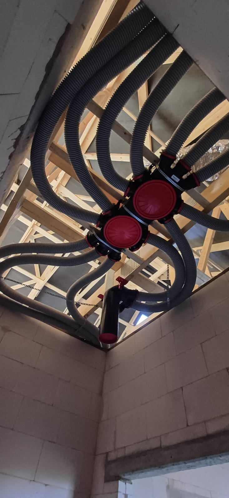 Rozprowadzenie szarych rur wentylacyjnych z czerwonymi elementami w surowym wnętrzu budynku. Widoczna konstrukcja drewniana stropu.