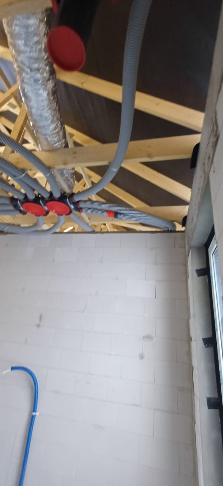 Szare rury wentylacyjne z czerwonymi elementami łączącymi, poprowadzone pod drewnianą konstrukcją dachu w surowym wnętrzu budynku z oknem.