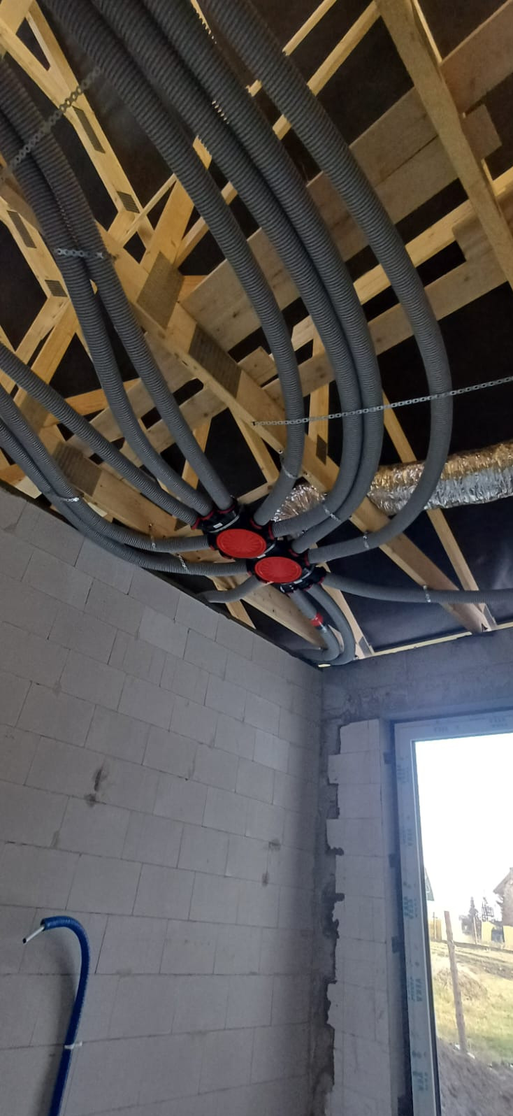 Szare rury wentylacyjne przymocowane do drewnianej konstrukcji sufitu w surowym wnętrzu. Widoczne czerwone elementy rozdzielcze i fragment ściany z oknem.