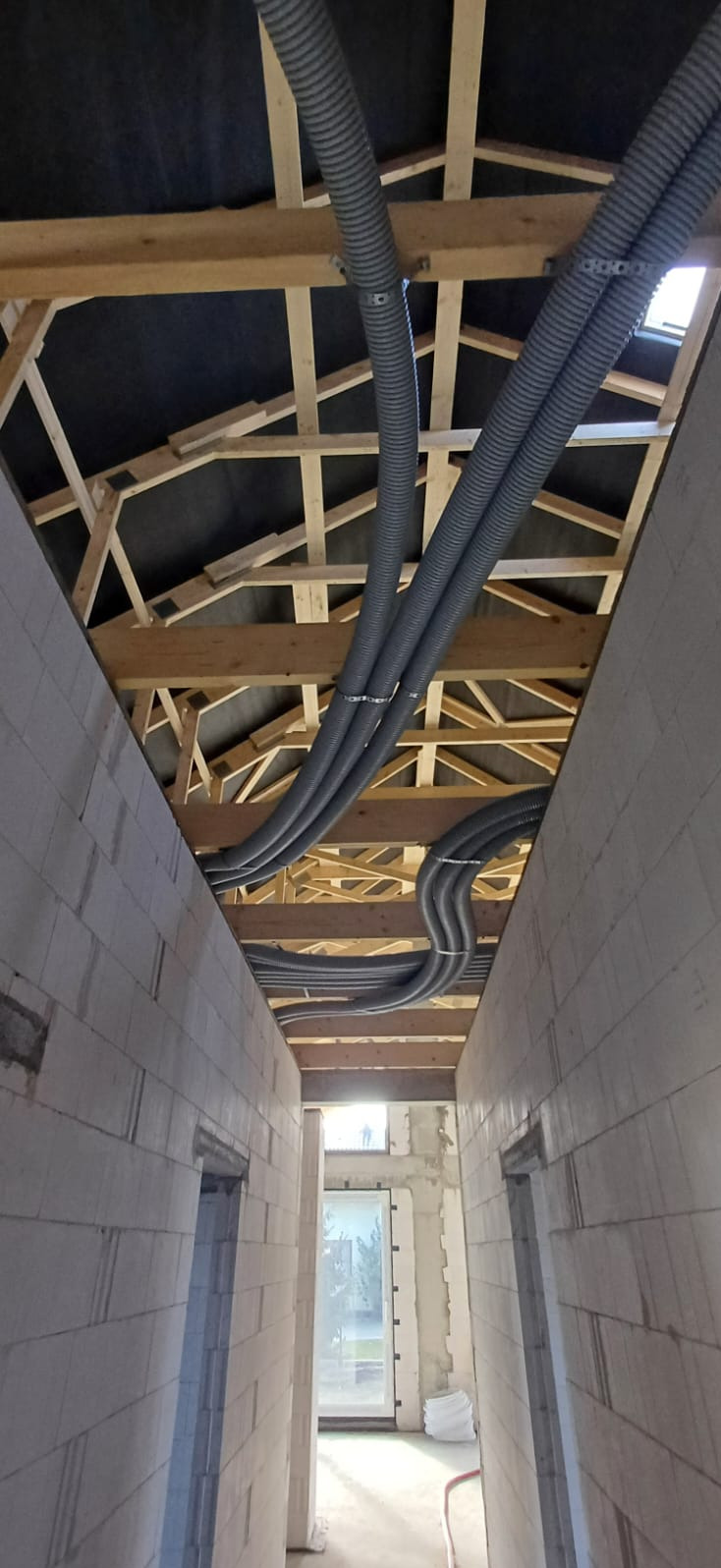 Szare, karbowane rury wentylacyjne pod drewnianą konstrukcją dachu w surowym wnętrzu budynku. Widoczne ściany z bloczków i otwór okienny w tle.