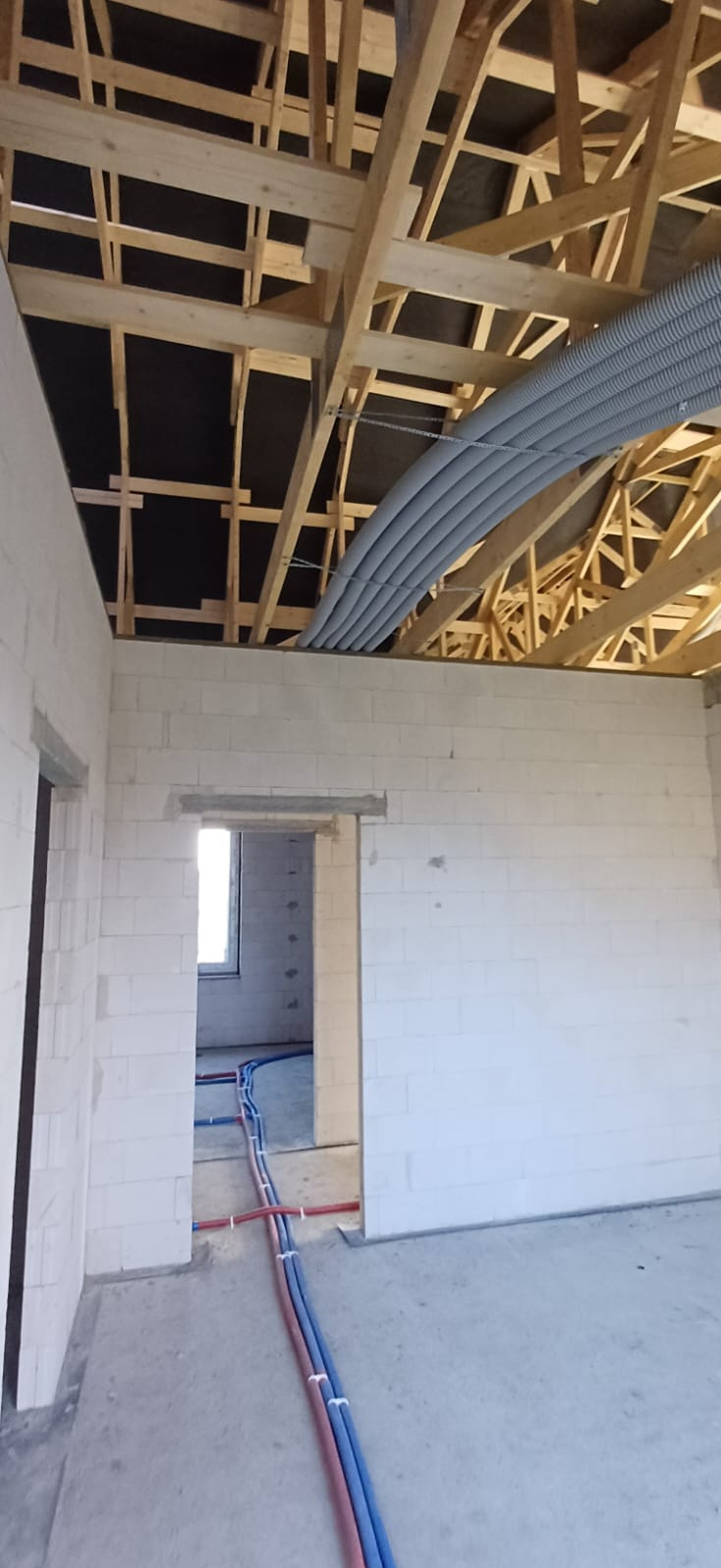 Instalacja rekuperacji: szare rury wentylacyjne pod drewnianym stropem, instalacja hydrauliczna na podłodze w surowym wnętrzu budynku.